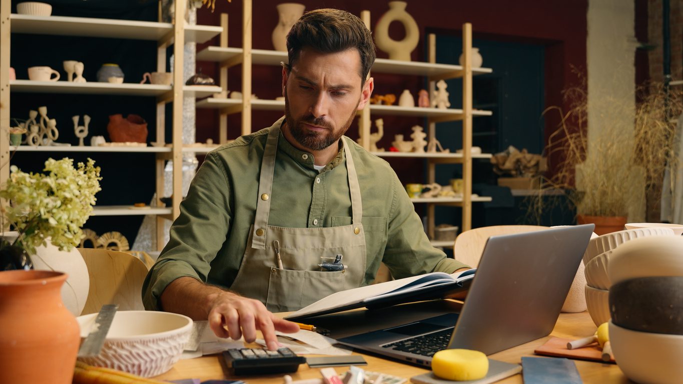 Serious frowned Caucasian man male small business owner businessman potter craftsman counting expense payment calculator taxes working laptop look notebook writing accounting indoors pottery workshop